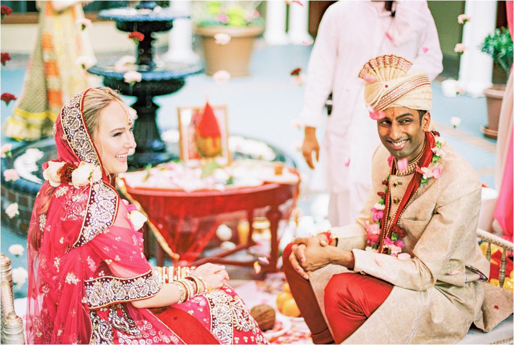 An Indian wedding ceremony in Charleston. The bride is wearing a pink saree and the groom is wearing a red and gold sherwani and they are smiling as guests throw flower petals on them.
