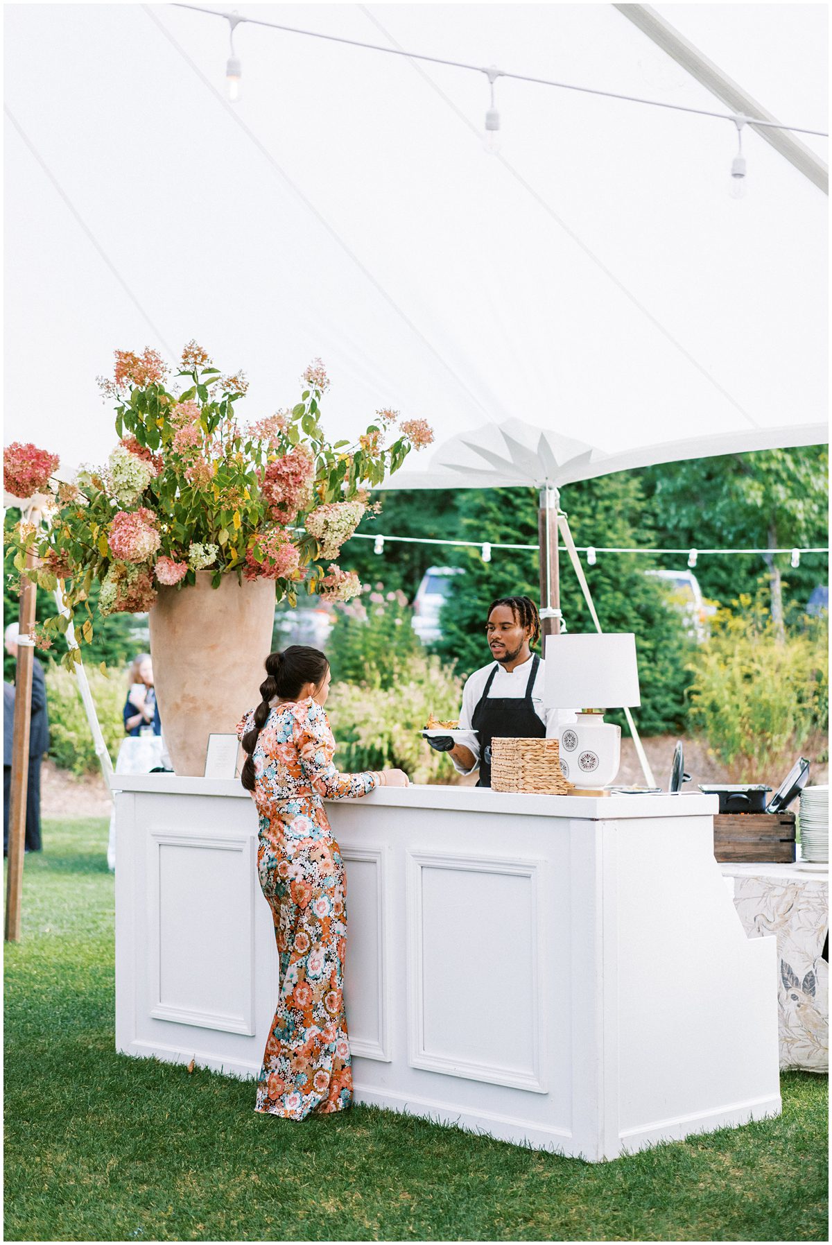 Candid wedding image of a well dressed guest in a colorful floor length floral dress ordering a drink at a white bar at a tented wedding reception in Highlands NC at the exclusive Mountaintop Golf and Lake Club