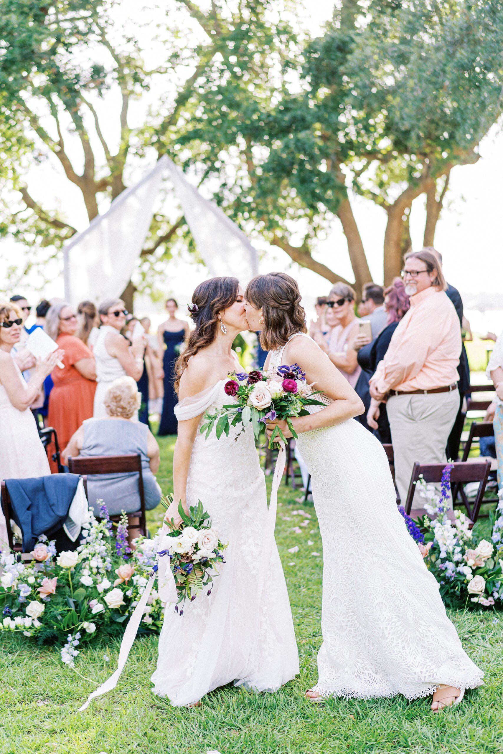 candid moment of two stylish brides kissing at the end of the ceremony aisle as guests look on at their wedding on the front lawn of Lowndes Grove in Charleston SC